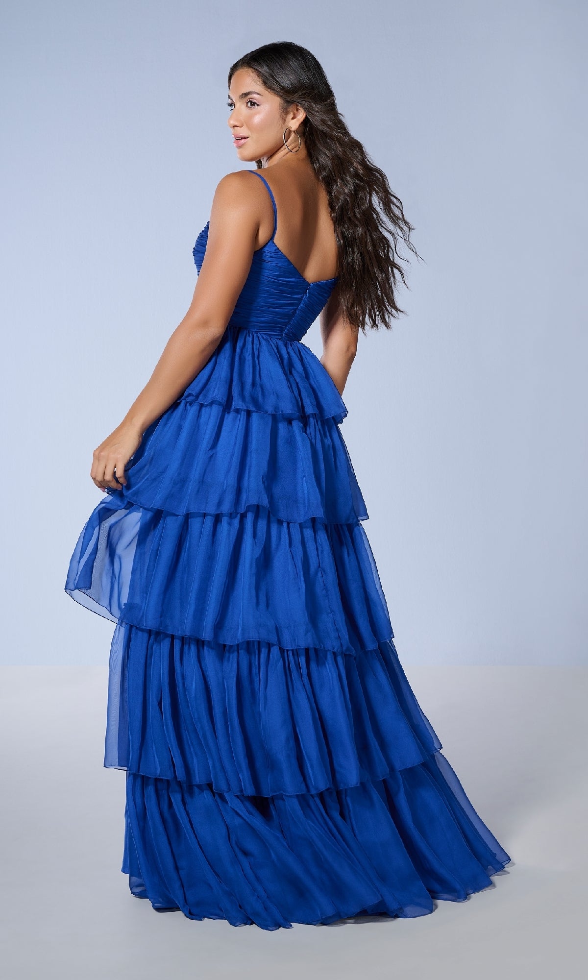 A woman in the Tiffany 16241 long prom dress—royal blue with a tiered, floor-length A-line ruffled skirt and spaghetti straps—looks over her shoulder against a plain light blue background.